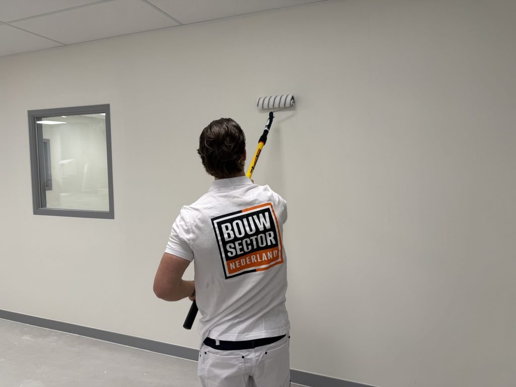 Schilder van Bouwsector Nederland zet een witte wand in de verf naast een binnenraam met grijs kozijn