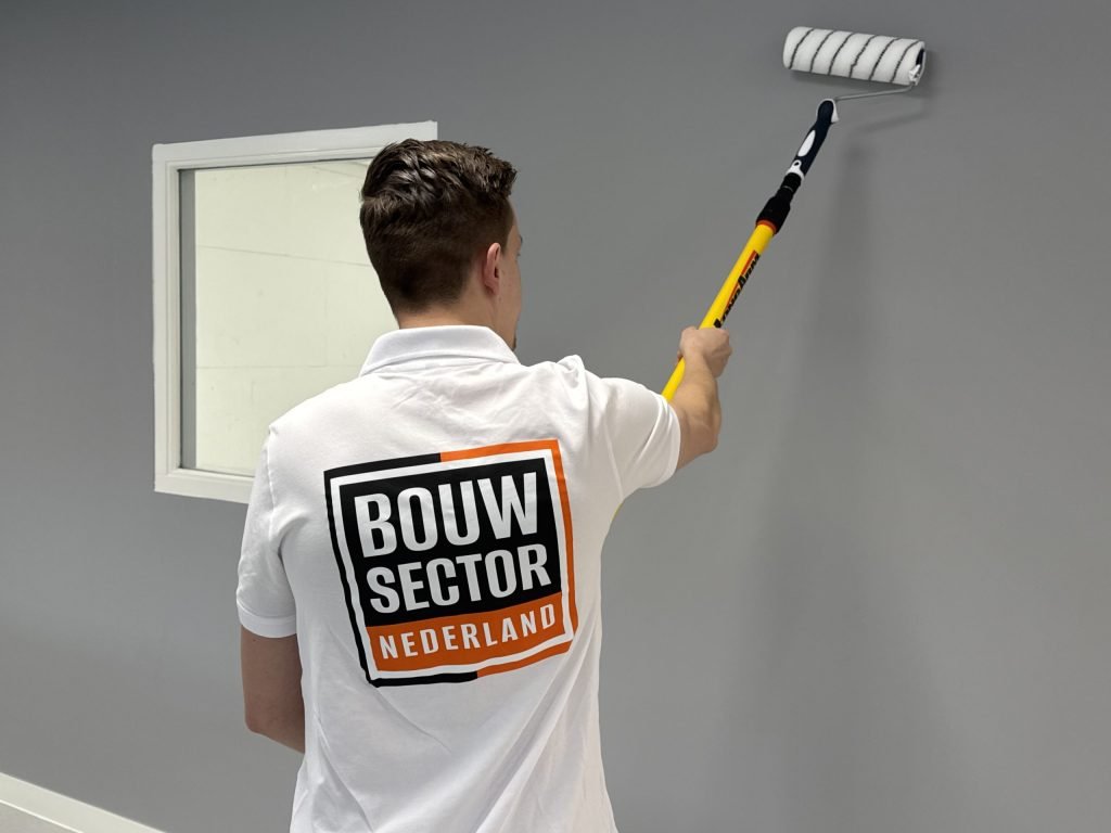 Schilder in een Bouwsector Nederland-shirt rolt een grijze muur naast een wit binnenraam