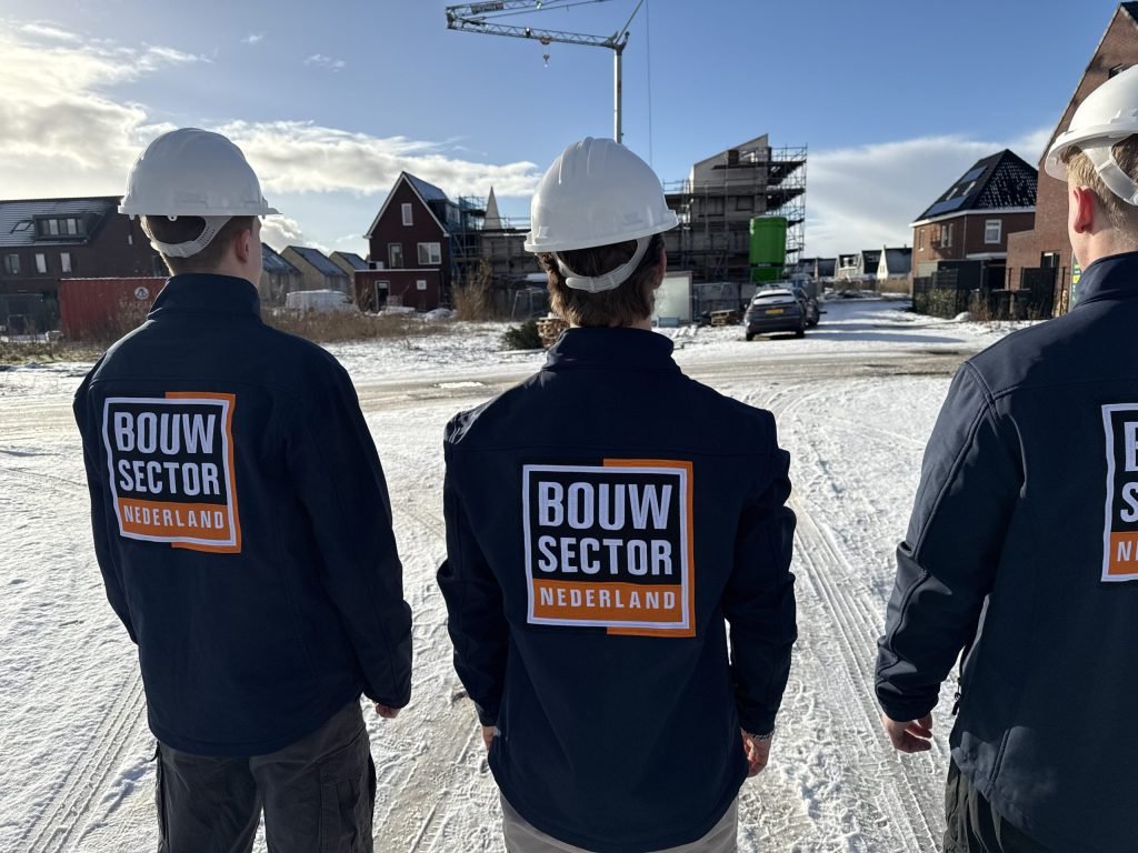 Drie vakmensen van Bouwsector Nederland in donkerblauwe werkjas met witte helm, op de rug gezien, bij een nieuwbouwwijk in de sneeuw